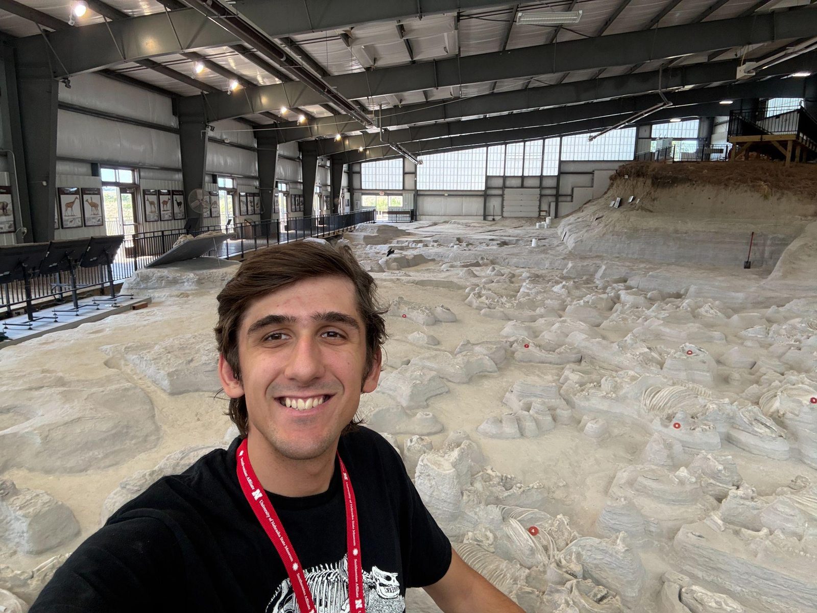 Luke inside the Hubbard Rhino Barn at Ashfall Fossil Beds, the ash bed behind him with numbered skeletons still lying where they were buried