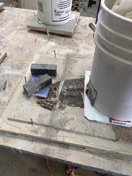 A fish plate sitting in a plywood form sealed around the edge with a bead of caulk, stone blocks and a bucket weighting pieces flat