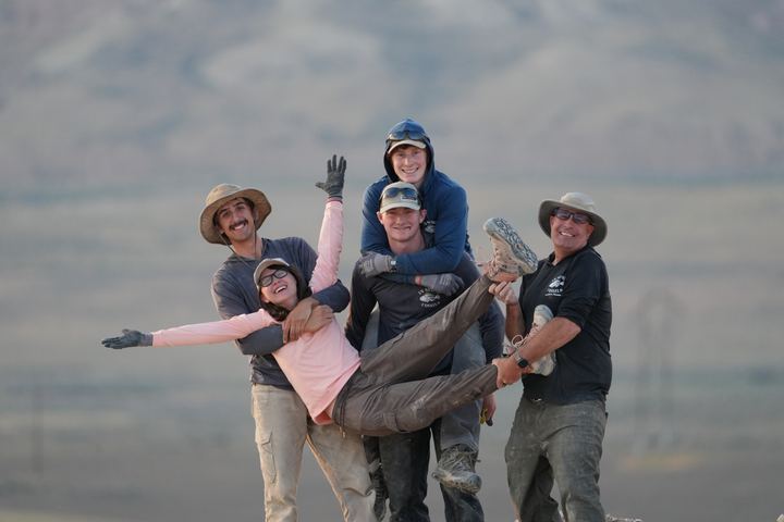 Five quarry crew members posing at the end of a day, one being carried by the others, dry hills behind them